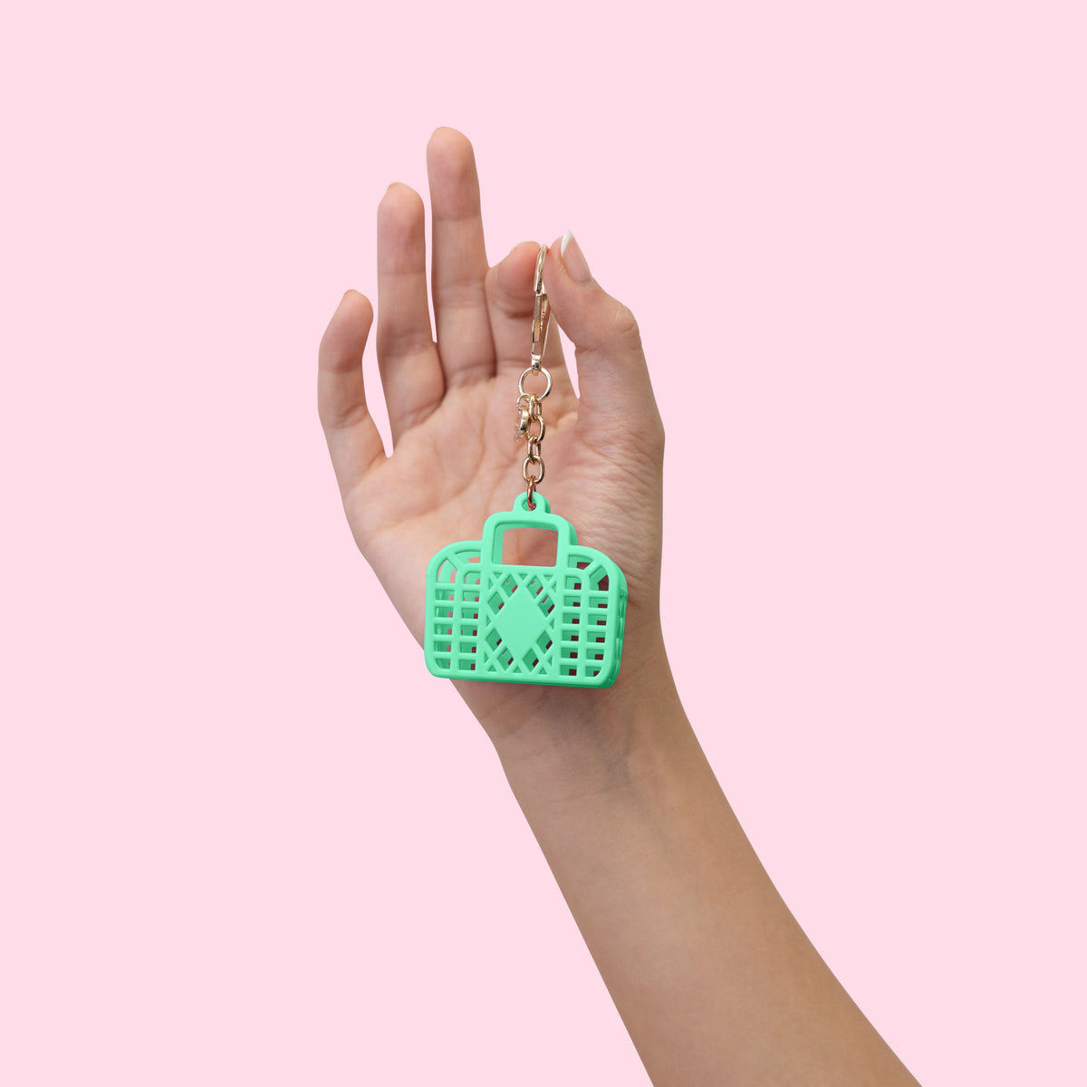 A hand holding a mint green plastic camera-shaped keychain with a gold metal chain against a solid light pink background.
