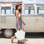 A woman wearing a brown wide-brim hat and blue denim overalls walks past a vintage cream-colored Volkswagen camper van, holding a white woven tote bag.